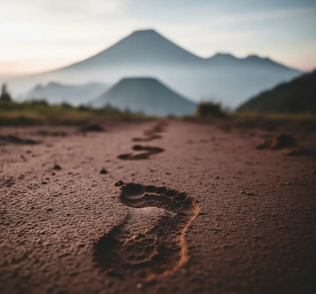 Jejak kaki di tanah mengarah ke pemandangan pegunungan, melambangkan perjalanan dan perenungan tentang legenda